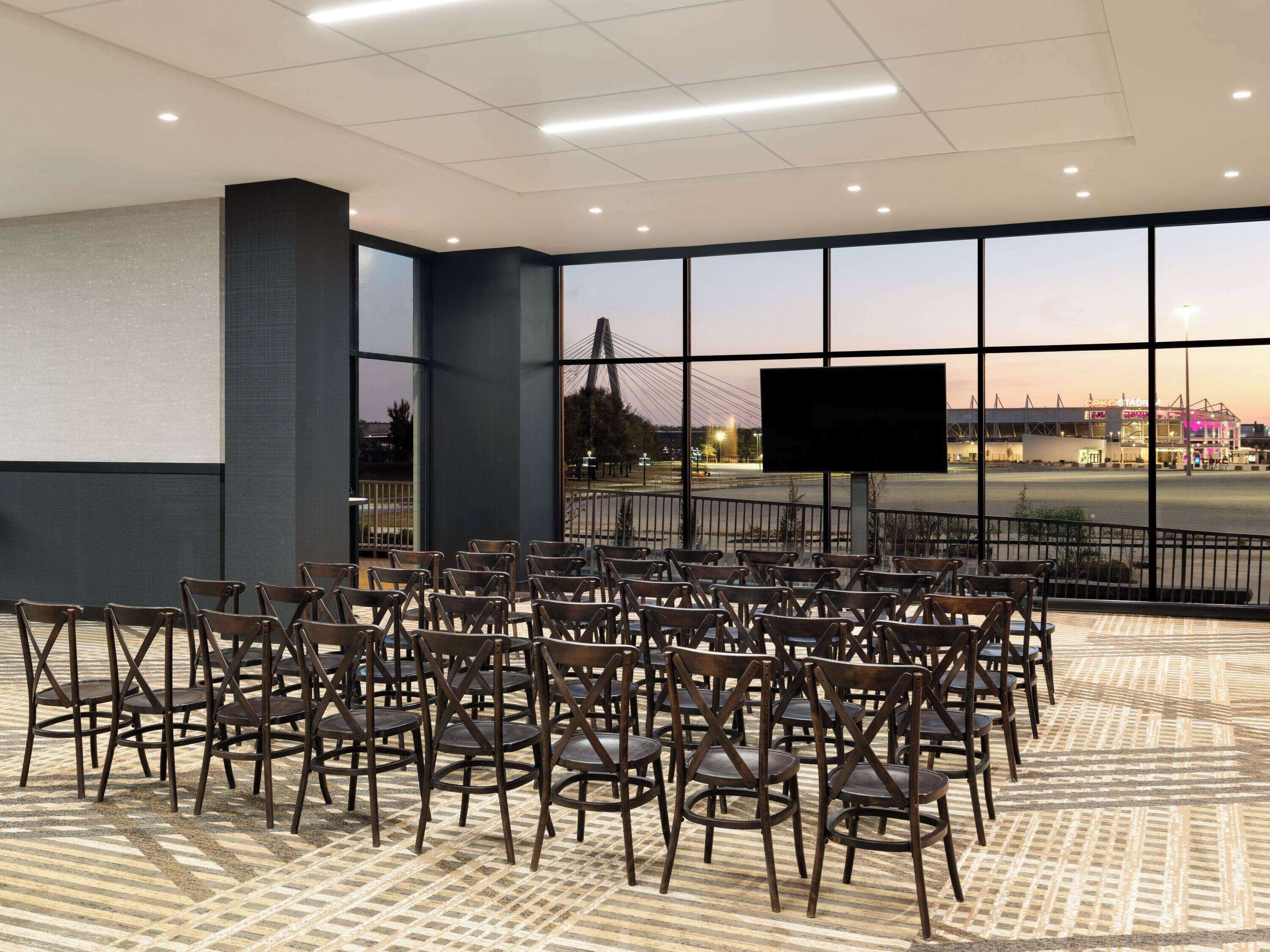A conference room with rows of wooden chairs, a large screen, and a view of a stadium.
