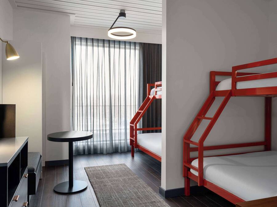 A modern hotel room with a bunk bed, a desk, a chair, a round table, and a large window with curtains.