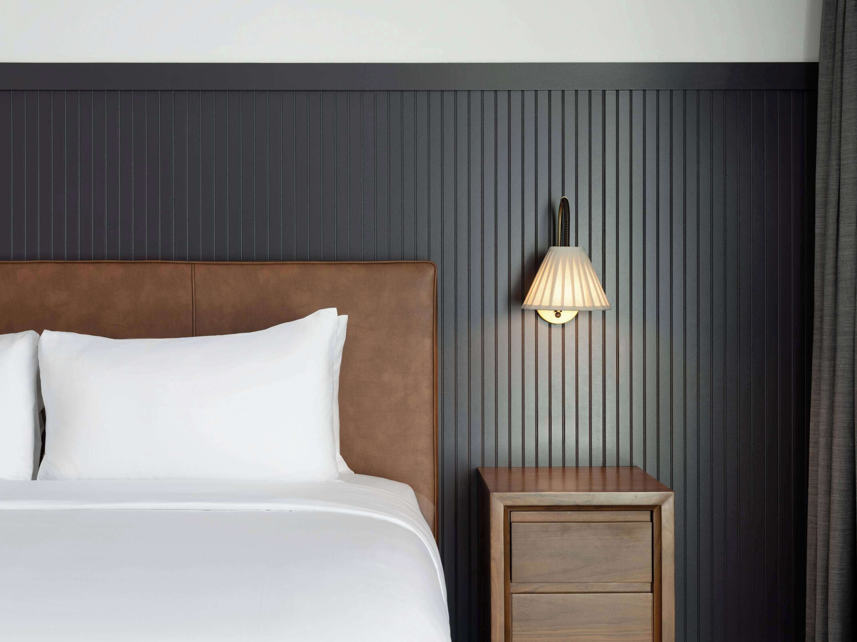 Un lit bien fait, avec du linge de lit blanc et une tête de lit en bois, une petite table de chevet en bois avec des tiroirs, et une lampe fixée au mur, le tout sur un fond de mur sombre à panneaux verticaux.