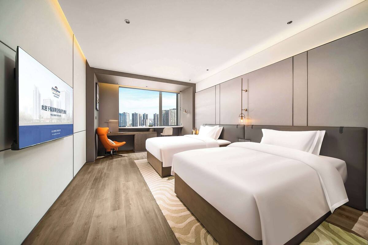 Habitación de hotel con dos camas, un escritorio, una silla y un ventanal con vista a la ciudad.