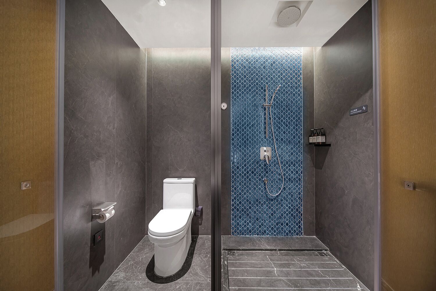 A modern bathroom with a toilet, a shower with a blue patterned wall, and a small shelf with toiletries.