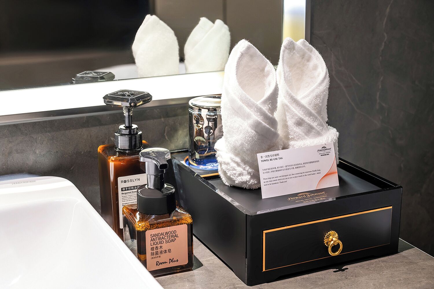 Un baño de hotel con un dispensador de jabón negro, una botella de jabón antibacteriano de sándalo, un dispensador de gel hidroalcohólico y un dispensador de toallas.