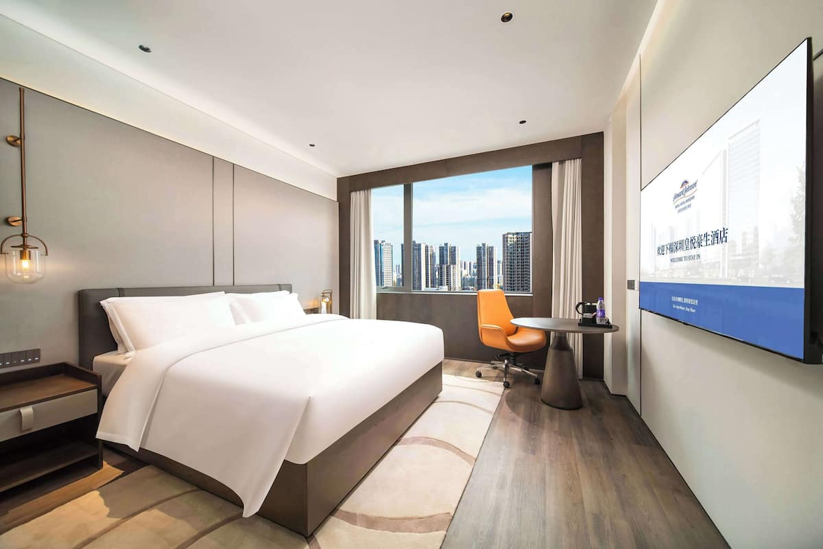 Una habitación de hotel moderna con una cama grande, un escritorio y una silla. Hay un ventanal con vistas a la ciudad.