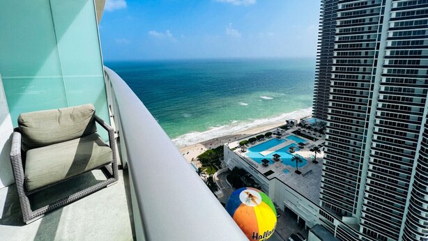 A balcony with a wicker chair overlooking a beach and a pool area. - Divine 2 Bedroom Apt at Lyfe Resort (Hollywood Beach)