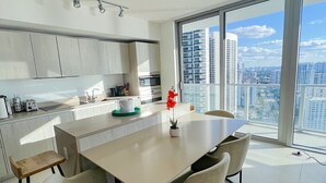 A modern kitchen with a large island, a dining area with chairs, and a view of the city through large windows. - Divine 2 Bedroom Apt at Lyfe Resort (Hollywood Beach)