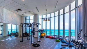 A gym with large windows overlooking the ocean, equipped with various exercise machines and weights. - Divine 2 Bedroom Apt at Lyfe Resort (Hollywood Beach)