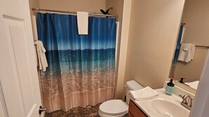 Bany amb cortina de dutxa amb escena de platja, un vàter, un lavabo amb dosificador de sabó i un mirall.