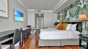 A hotel room with a bed, desk, and chair, a painting of a beach, and a tropical wallpaper. - Spectacular Bay View 1bd 2baths in Grove (Miami)