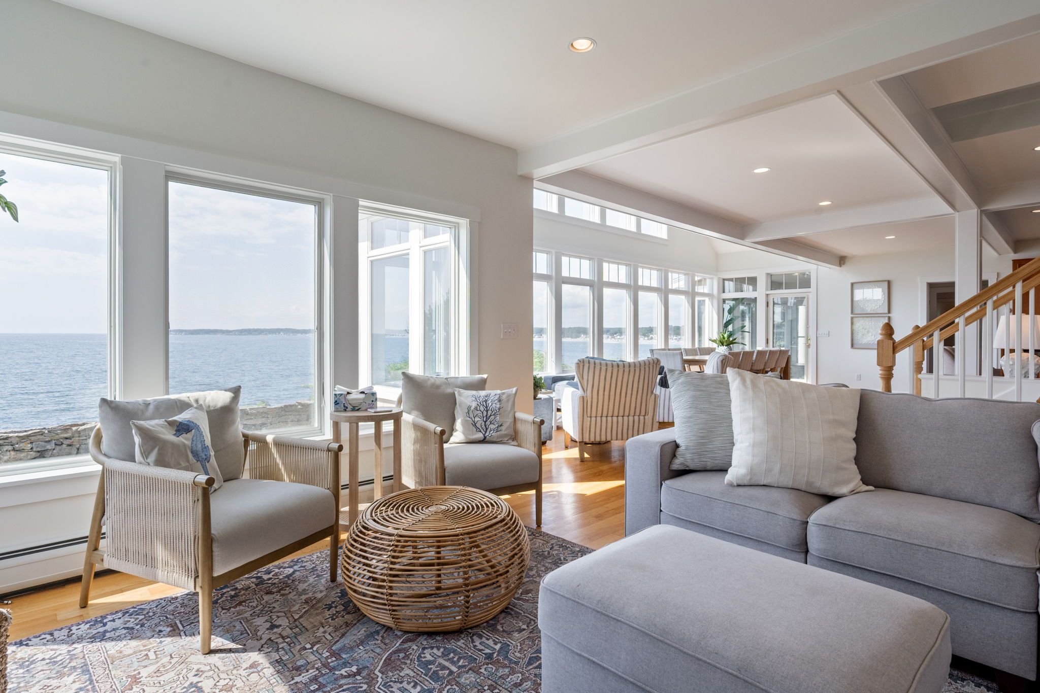 A spacious living room with a view of the sea, large windows, a grey sofa, and a wicker coffee table.