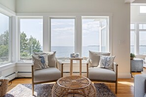 A room with two chairs facing a large window overlooking a body of water, a small table, and a woven floor mat. - Spacious York Home with Ocean Views & Hot Tub (York)