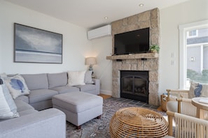A living room with a grey sectional sofa, a stone fireplace, a flat-screen TV mounted above the fireplace, and a large landscape painting on the wall. - Spacious York Home with Ocean Views & Hot Tub (York)