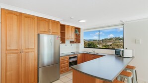 A modern kitchen with wooden cabinets, a stainless steel refrigerator, a microwave, and a large window offering a view of a hilly landscape.