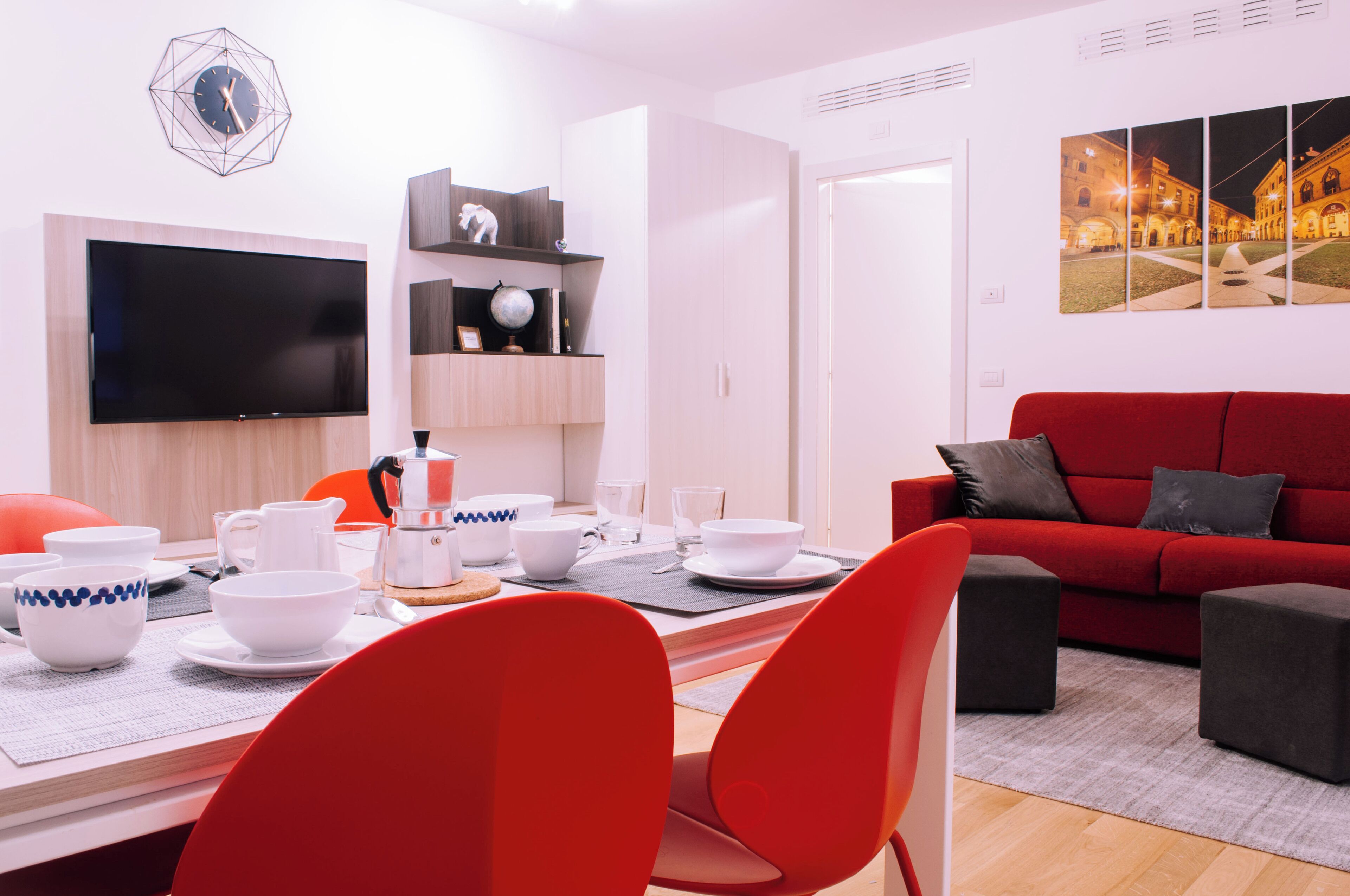 A modern living room with a red sofa, a dining table set with white dishes, and a flat-screen TV mounted on the wall.