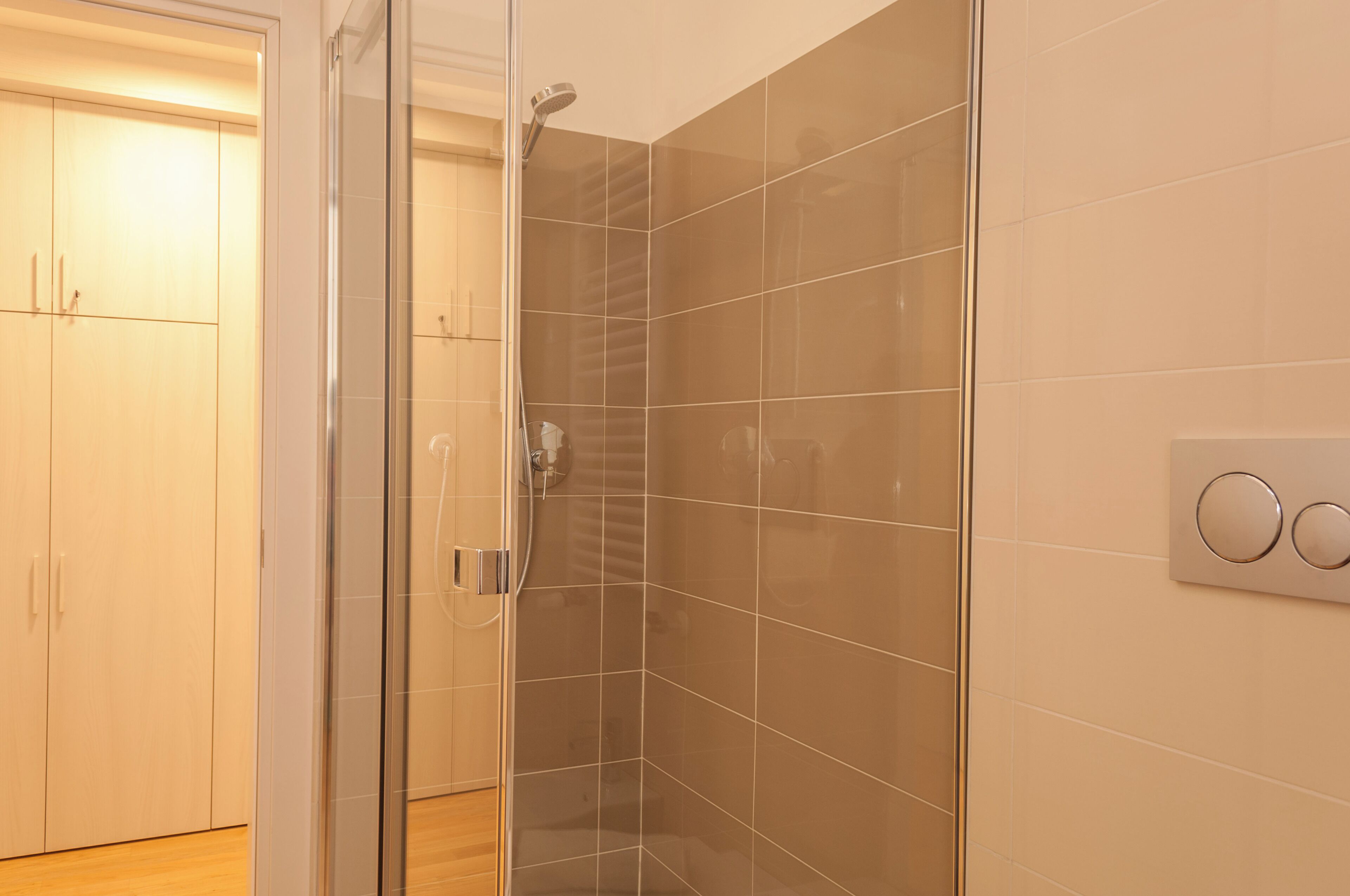 A modern bathroom with a glass shower enclosure, a white tiled wall, and a wooden floor.