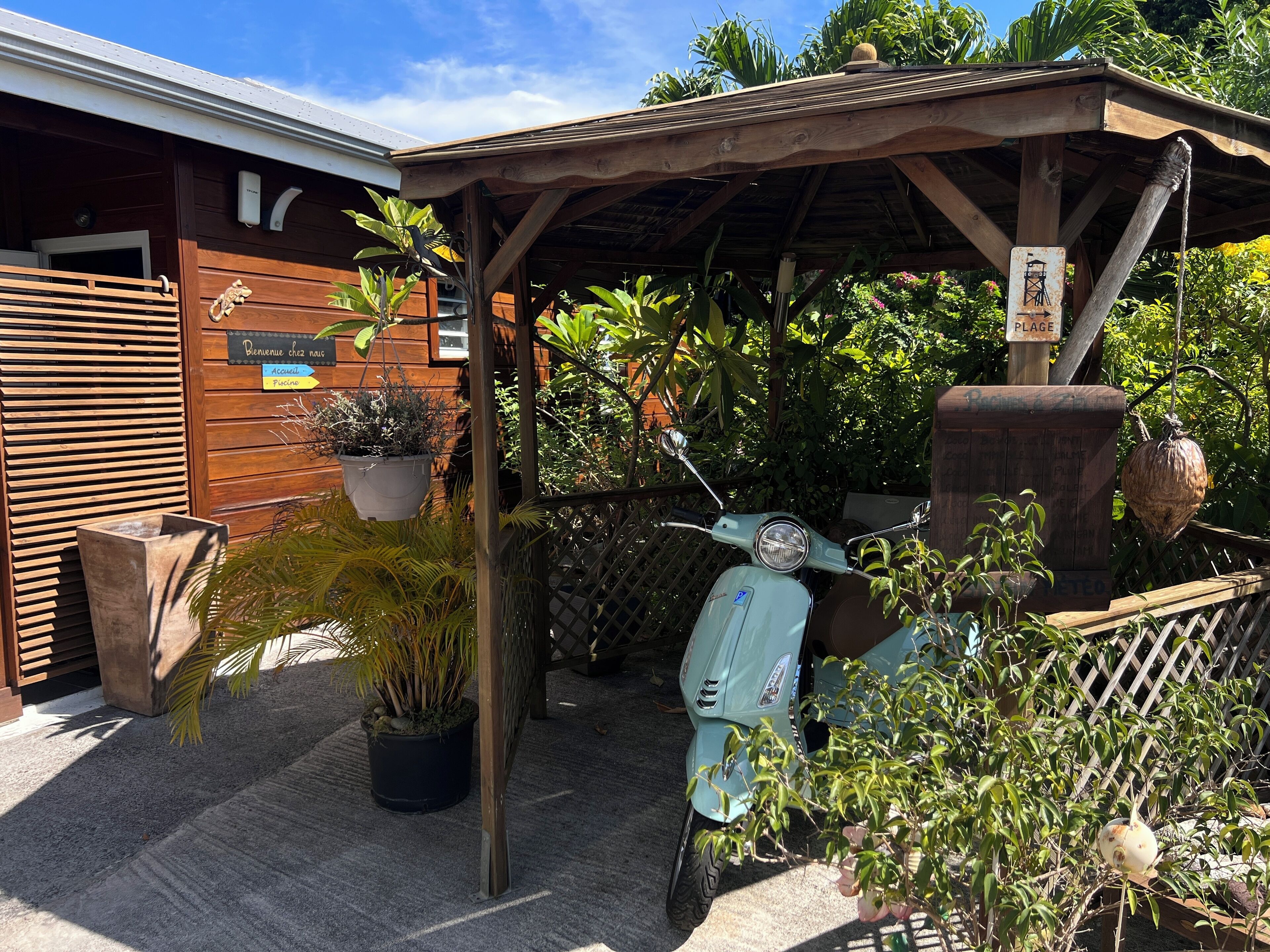 A covered outdoor seating area with a scooter parked underneath, surrounded by potted plants and a wooden fence.