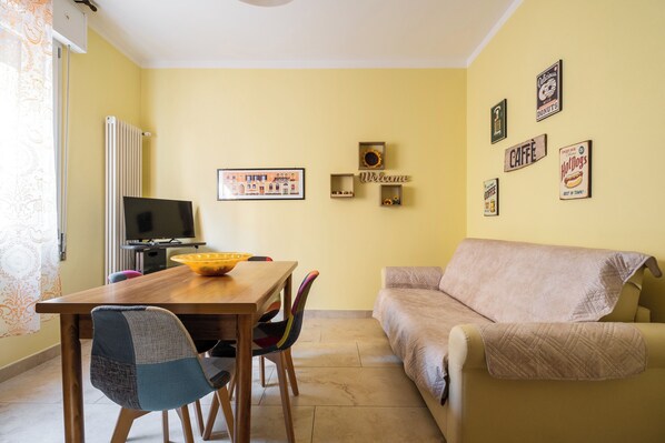A living room with a beige sofa, a wooden dining table with chairs, a TV, and wall decorations including framed pictures and signs. - San Donato Cozy Apartment by Wonderful Italy (Bologna)