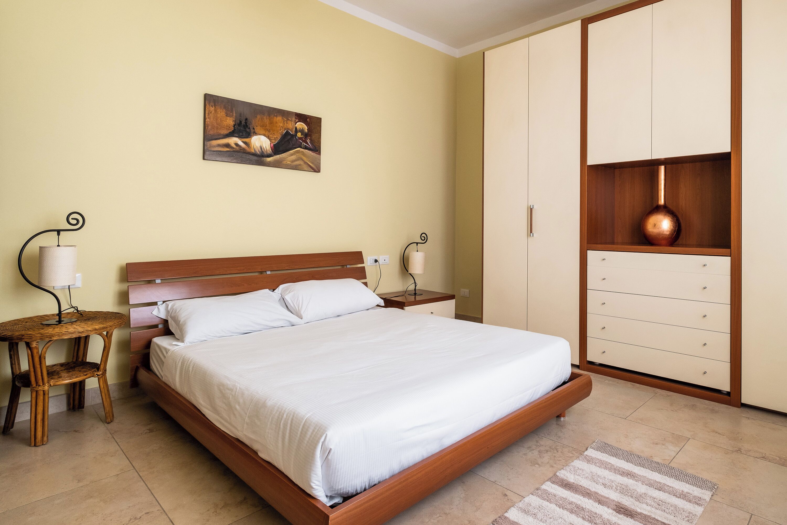 A bedroom with a wooden bed, a nightstand with a lamp, a painting on the wall, and a wardrobe with a copper vase.