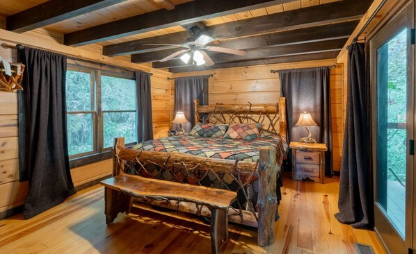 A log cabin bedroom with a wooden bed, a bench, a ceiling fan, and large windows.