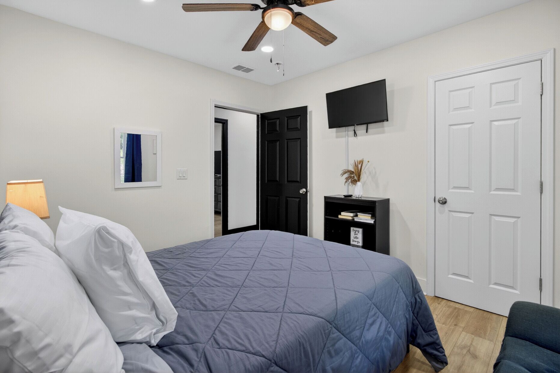 A bedroom with a bed, a ceiling fan, a TV, a door, and a small table with books.