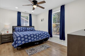 A bedroom with a bed, a ceiling fan, a window with curtains, a bedside table with a lamp, and a rug.