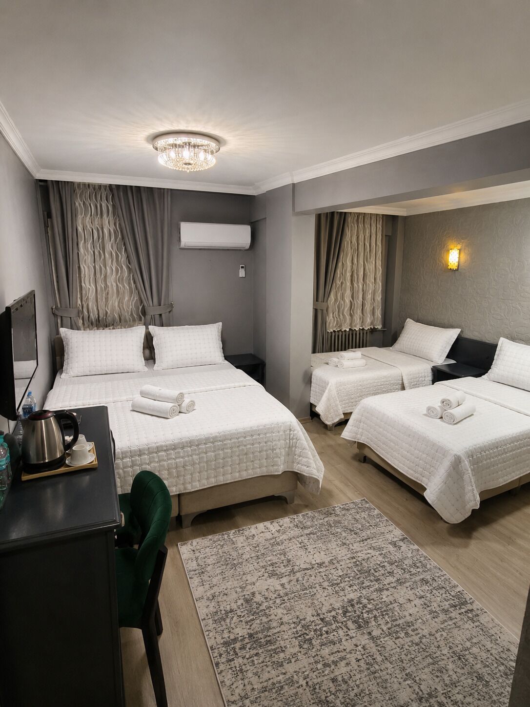 A hotel room with two beds, a desk with a kettle, a chair, and a large window with curtains.