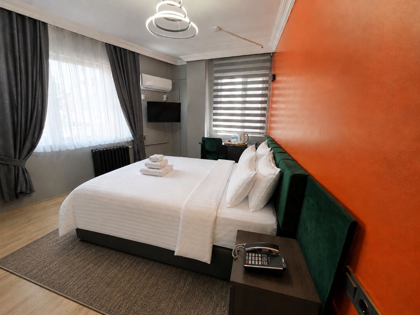 A hotel room with a bed, a bedside telephone, a radiator, and a window with blinds.