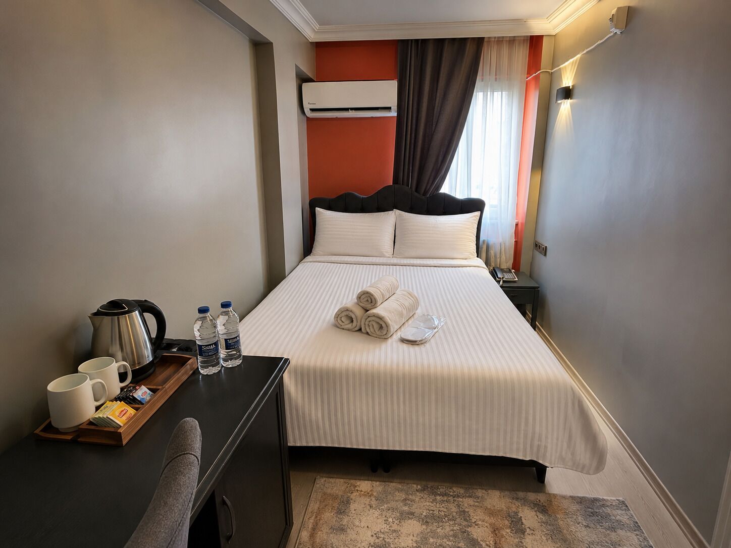 Habitació d'hotel amb llit, taula de nit i escriptori, on hi ha un búl, ampolles d'aigua i una safata amb gots i un llibre.