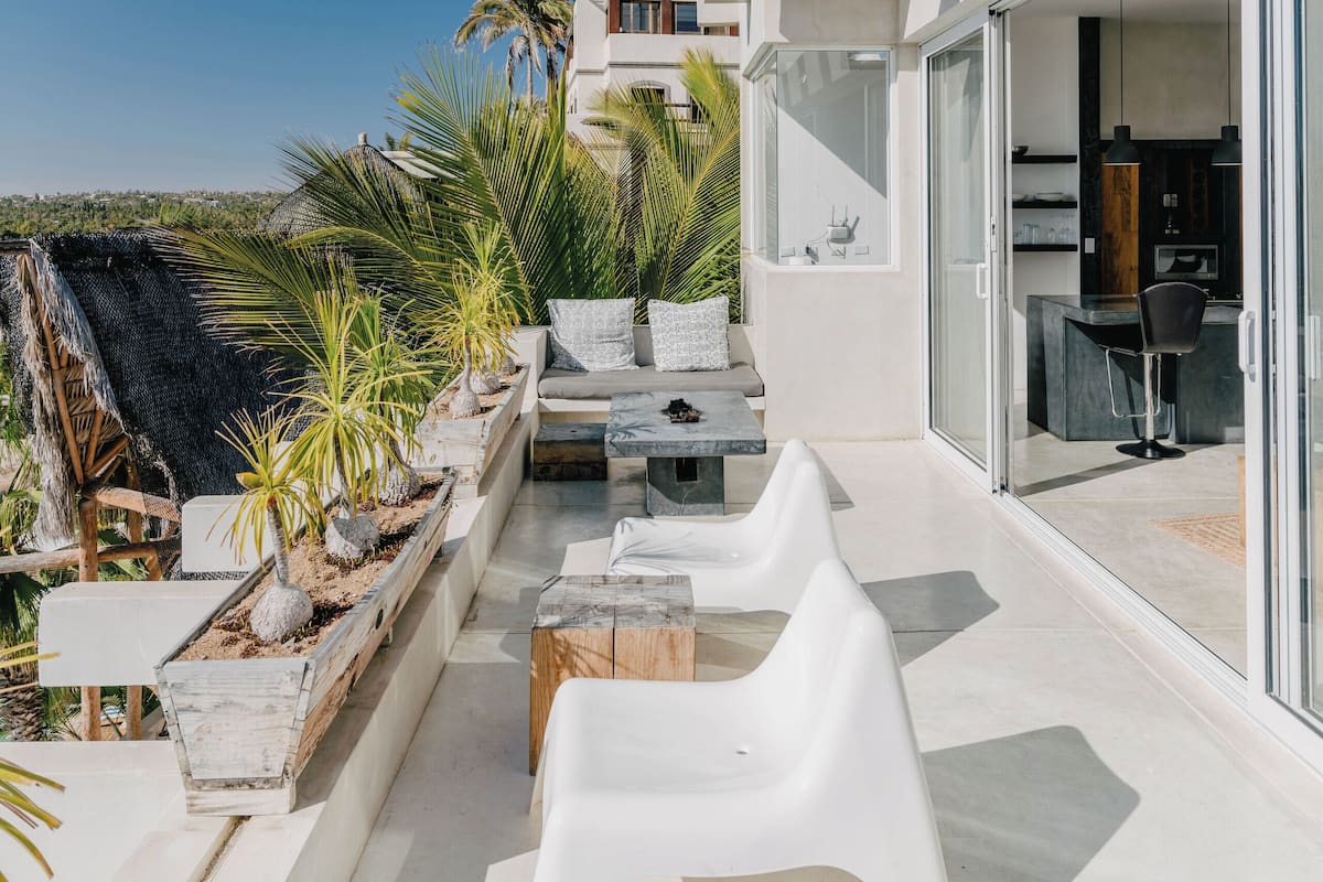 A modern outdoor patio with white furniture, palm plants, and a view of the surrounding landscape.