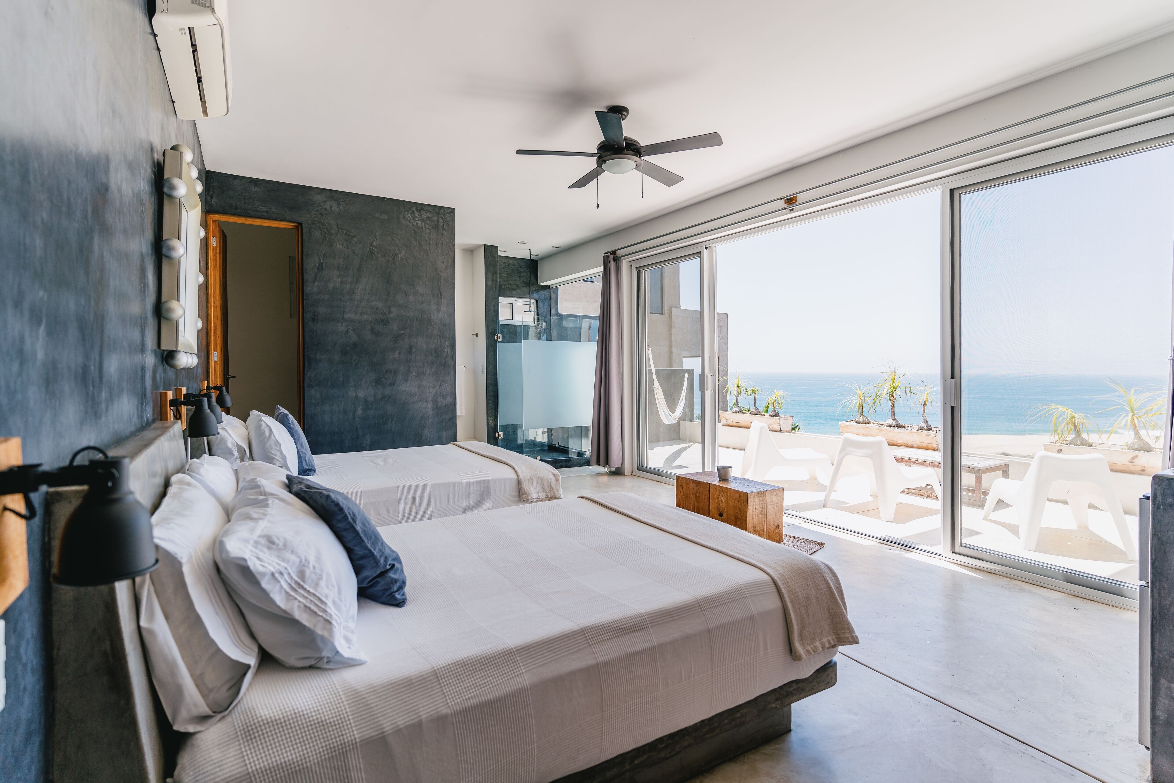 Una habitación de hotel moderna con dos camas, un ventilador de techo y un amplio ventanal corredero que da a un balcón con vista al mar.