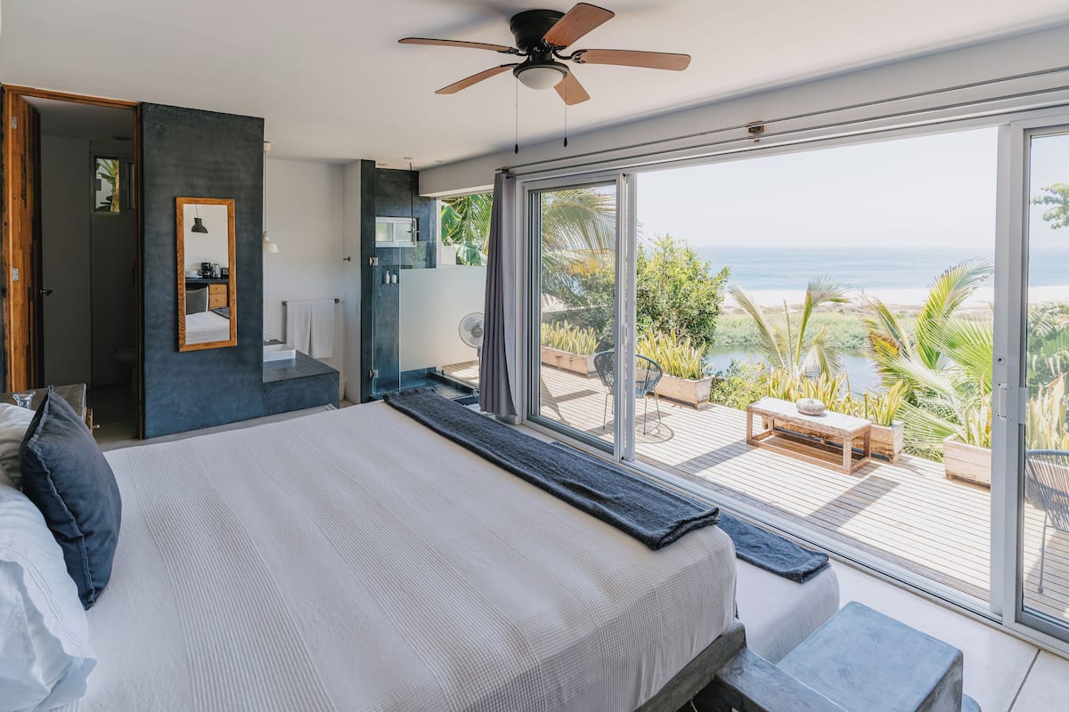 A modern bedroom with a large bed, a ceiling fan, and a view of the ocean through floor-to-ceiling windows.