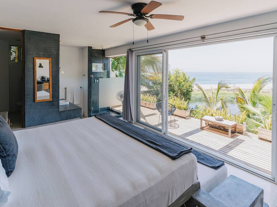 A modern bedroom with a large bed, a ceiling fan, and a view of the ocean through floor-to-ceiling windows.