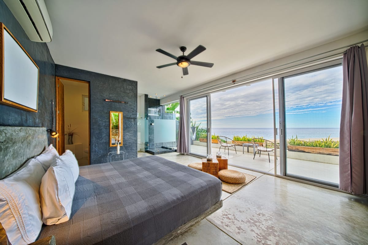 A modern bedroom with a large bed, a ceiling fan, and a view of the ocean through floor-to-ceiling windows.