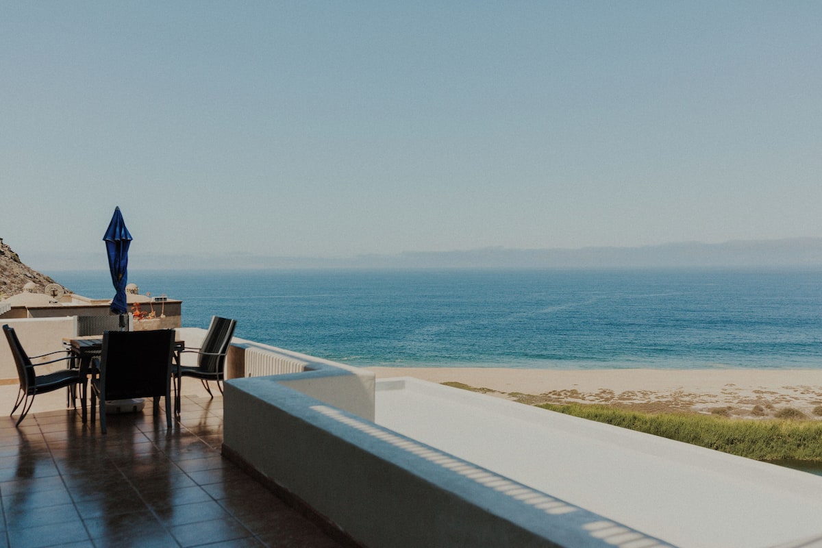 A terrace with a table and chairs overlooking a beach and the sea.