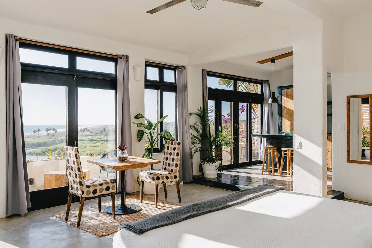 A spacious room with a large bed, a dining table with chairs, and a view of the outdoors through large windows.