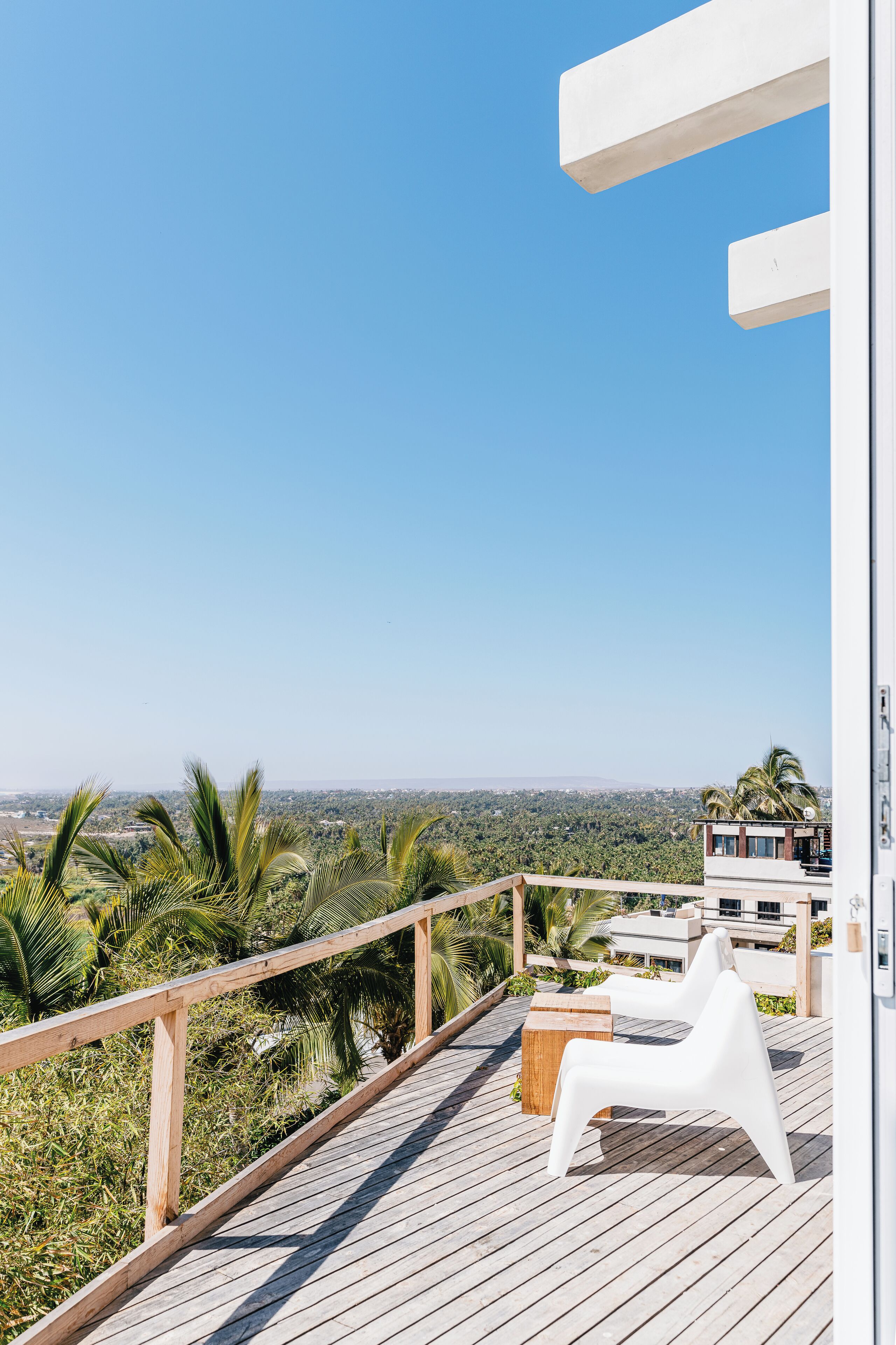 Una terraza de madera con una silla blanca y una mesa de madera, con vista a un paisaje urbano y palmeras.