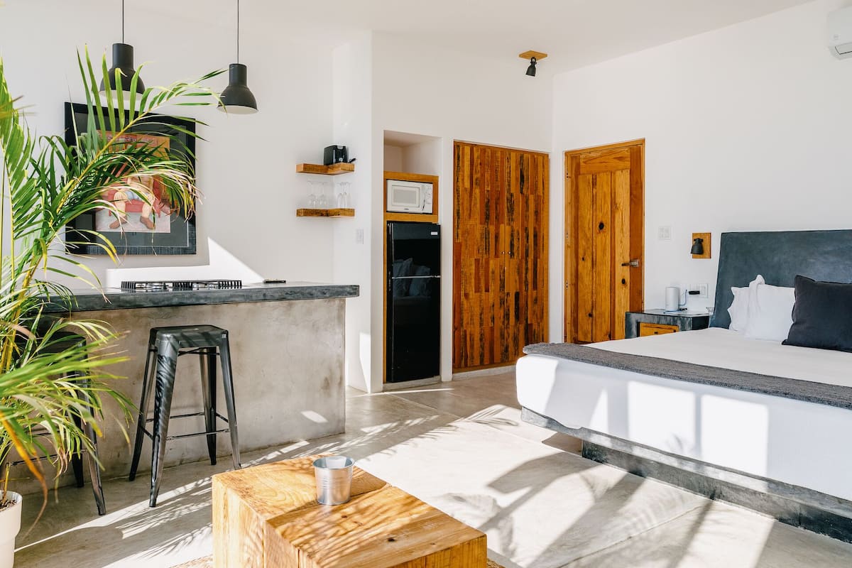 A modern hotel room with a large bed, a small kitchenette, a wooden wardrobe, and a dining area with a bar stool.