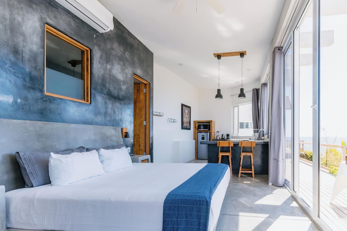 A modern bedroom with a large bed, a small kitchenette, and a balcony with a view.