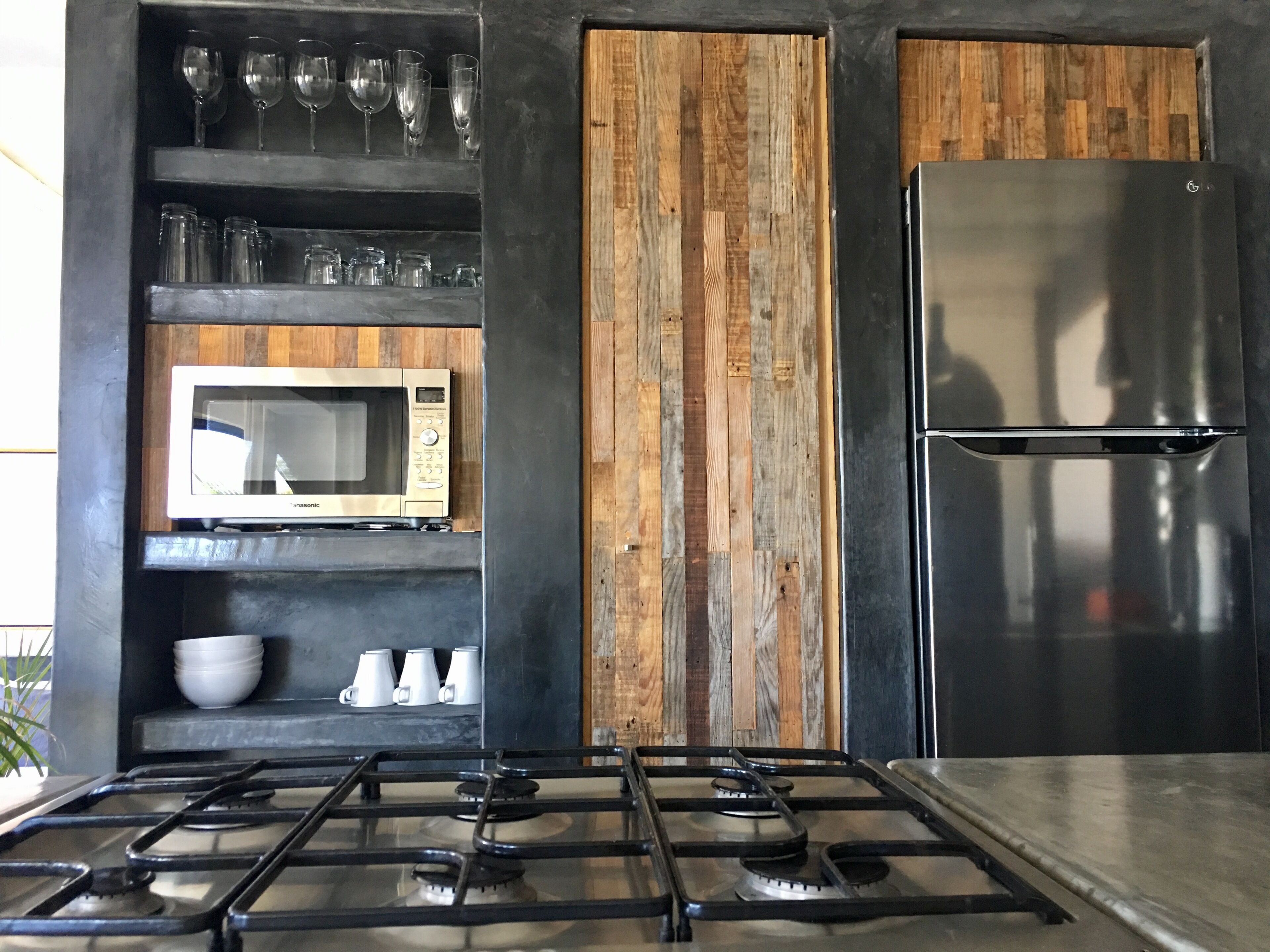 A kitchen with a gas stove, a microwave, and a stainless steel refrigerator. There are shelves with glassware and cups.