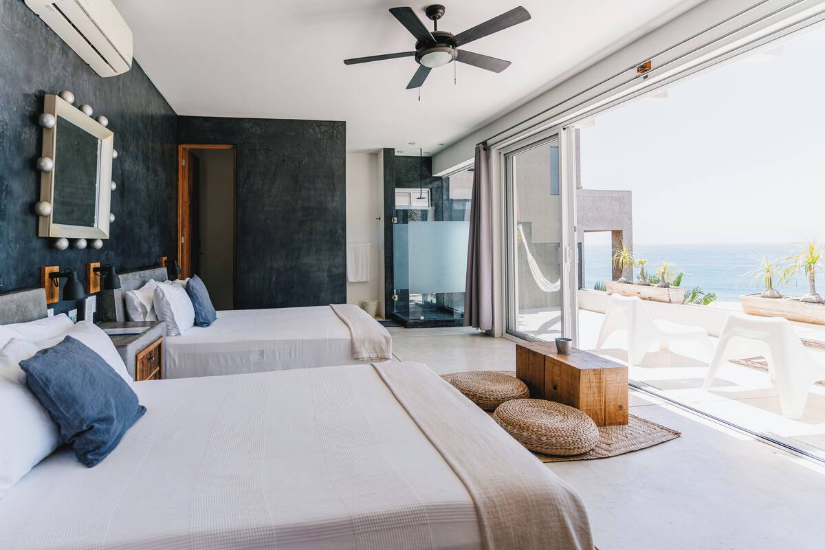 A modern hotel room with two beds, a ceiling fan, and a balcony with a view of the sea.