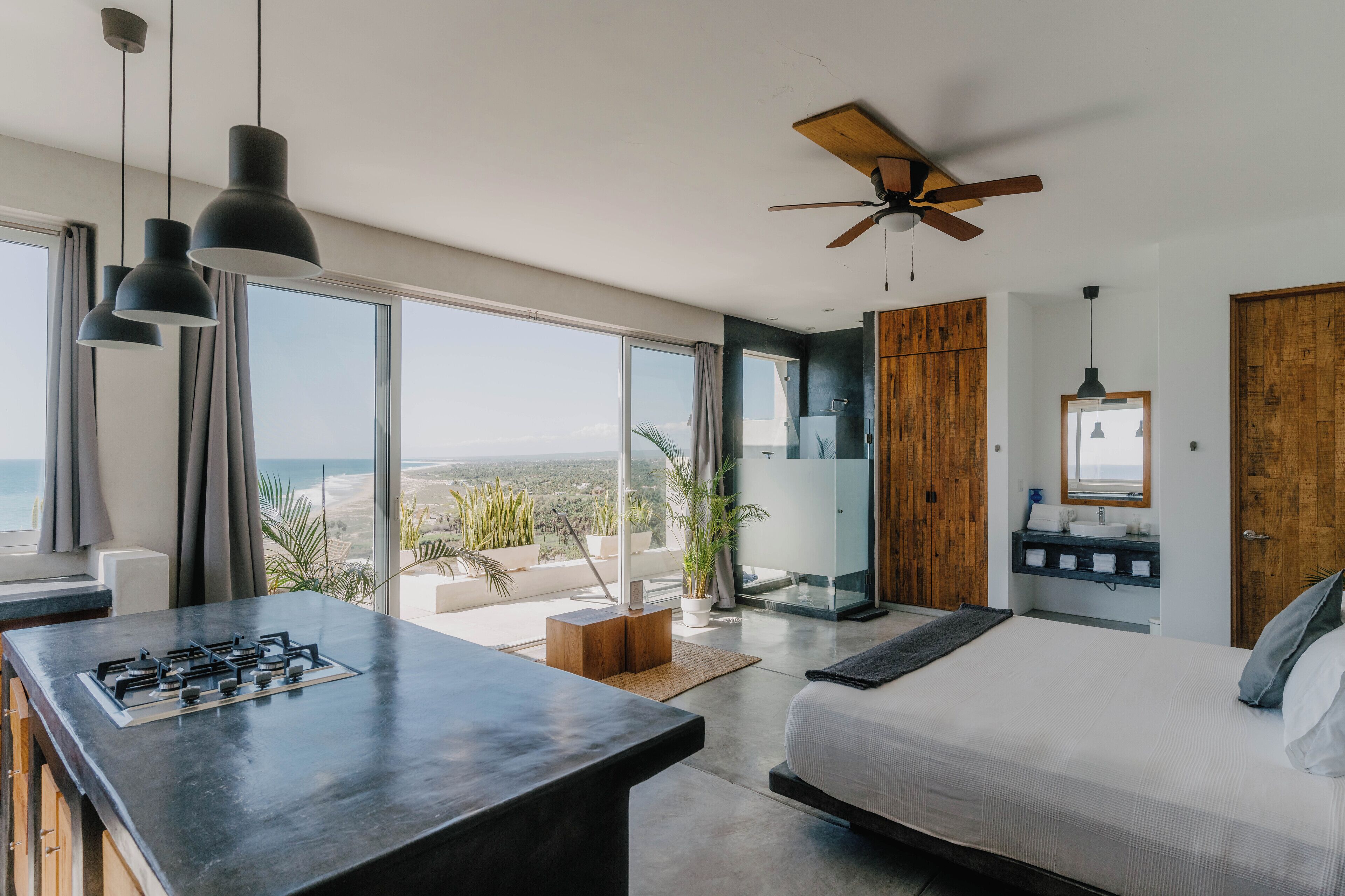 A modern bedroom with a large bed, a kitchen area with a gas stove, and a view of the ocean through large windows.