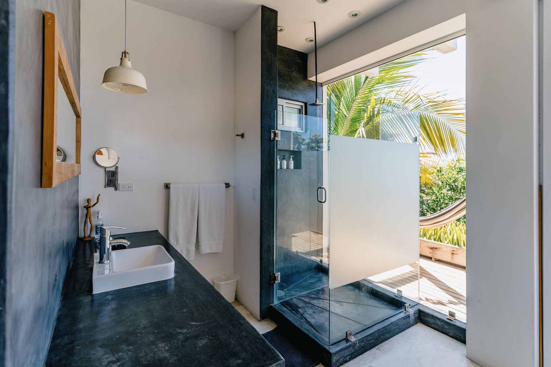 Un baño moderno con encimera negra, cabina de ducha con paredes de vidrio y vistas al exterior con una hamaca y palmeras.