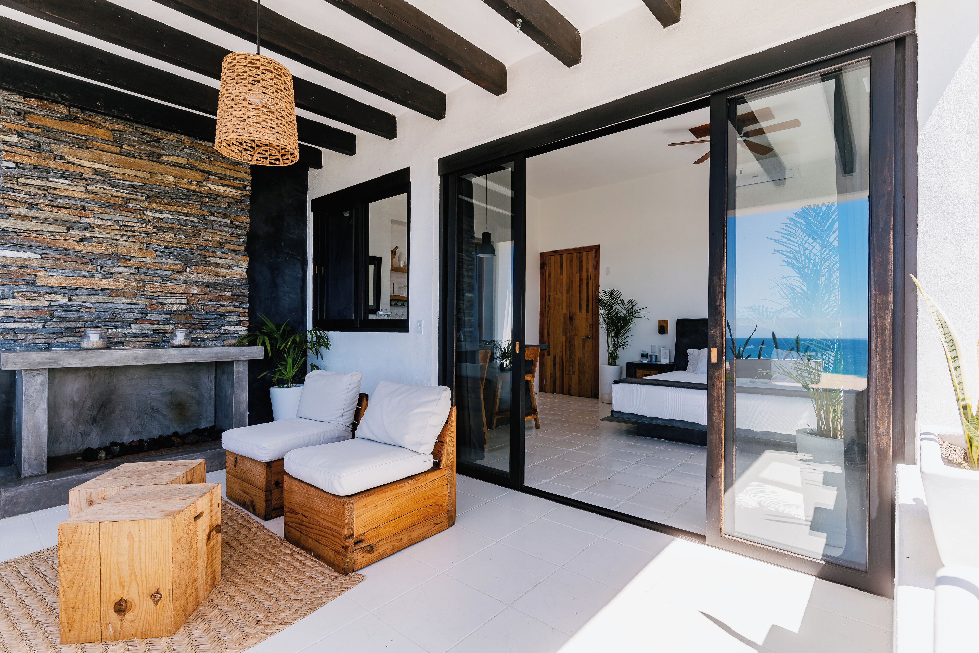 A modern outdoor living area with a stone wall, wooden furniture, and a glass door leading to a balcony with a view of the sea.