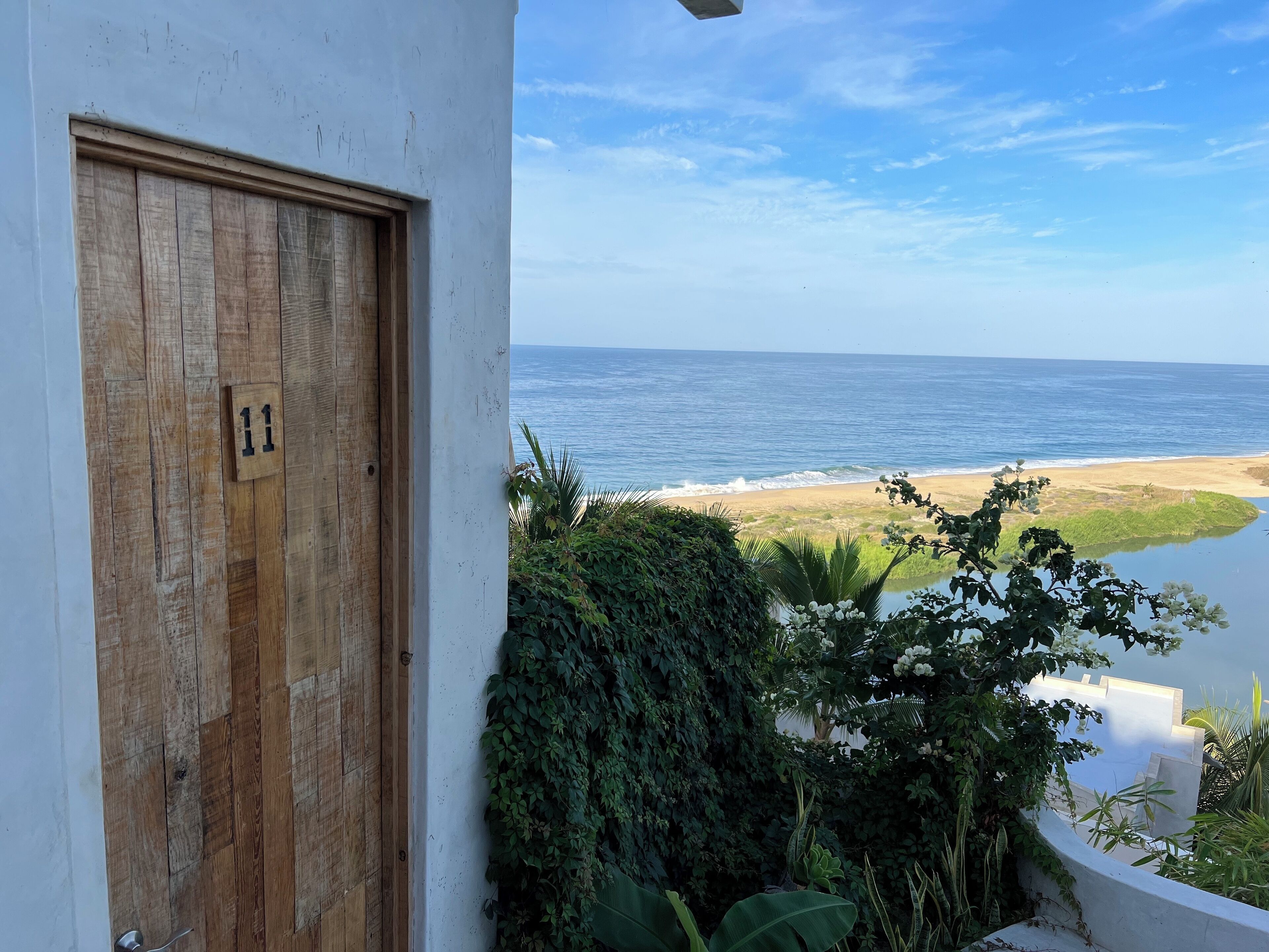 Una puerta de madera con una placa de número, una pared blanca y vista a la playa y al océano.