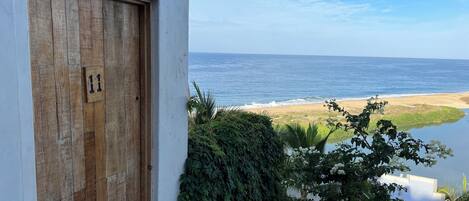 Una puerta de madera con una placa de número, una pared blanca y vista a la playa y al océano.