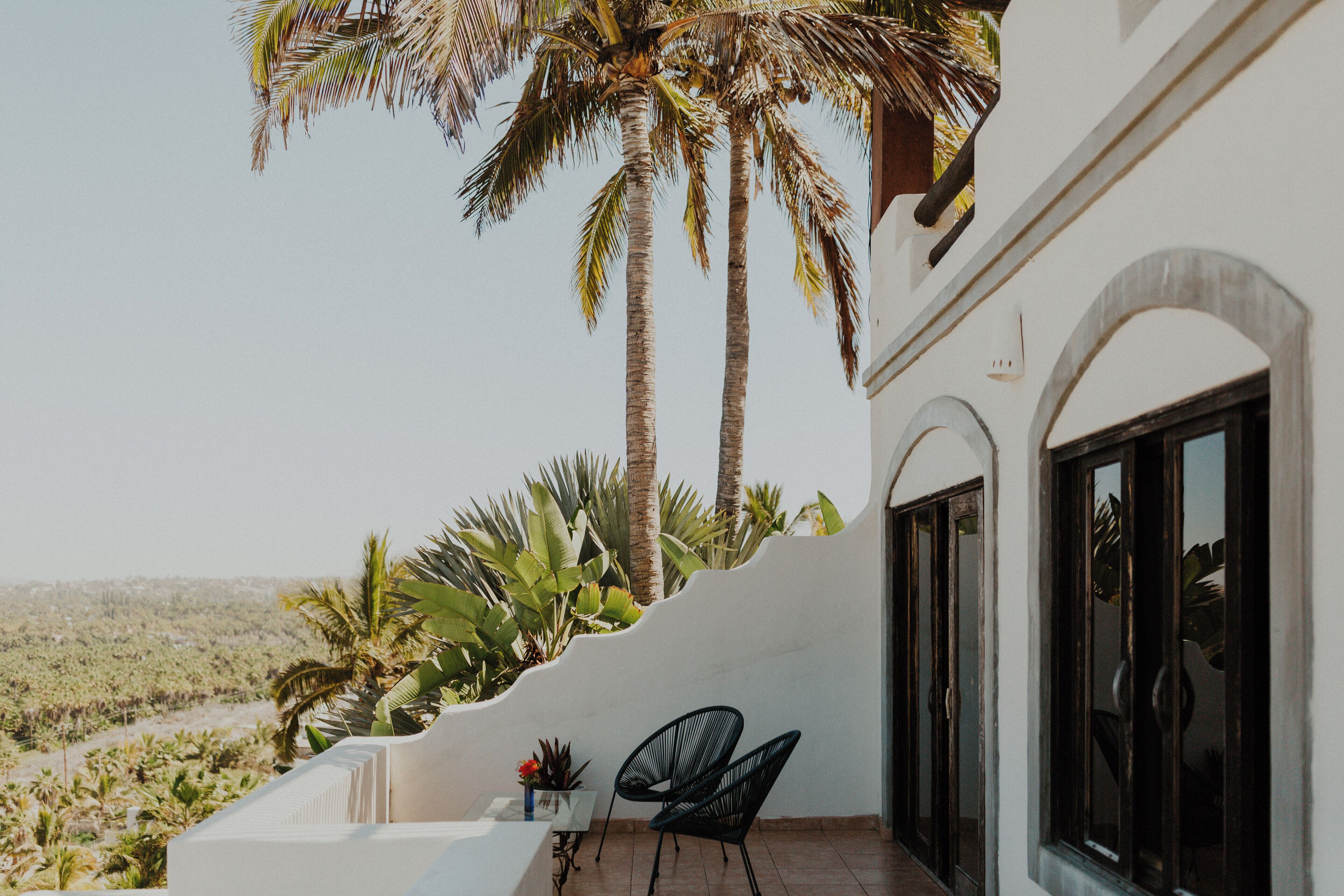 A balcony with a white wall, two black chairs, and a view of palm trees and a desert landscape.