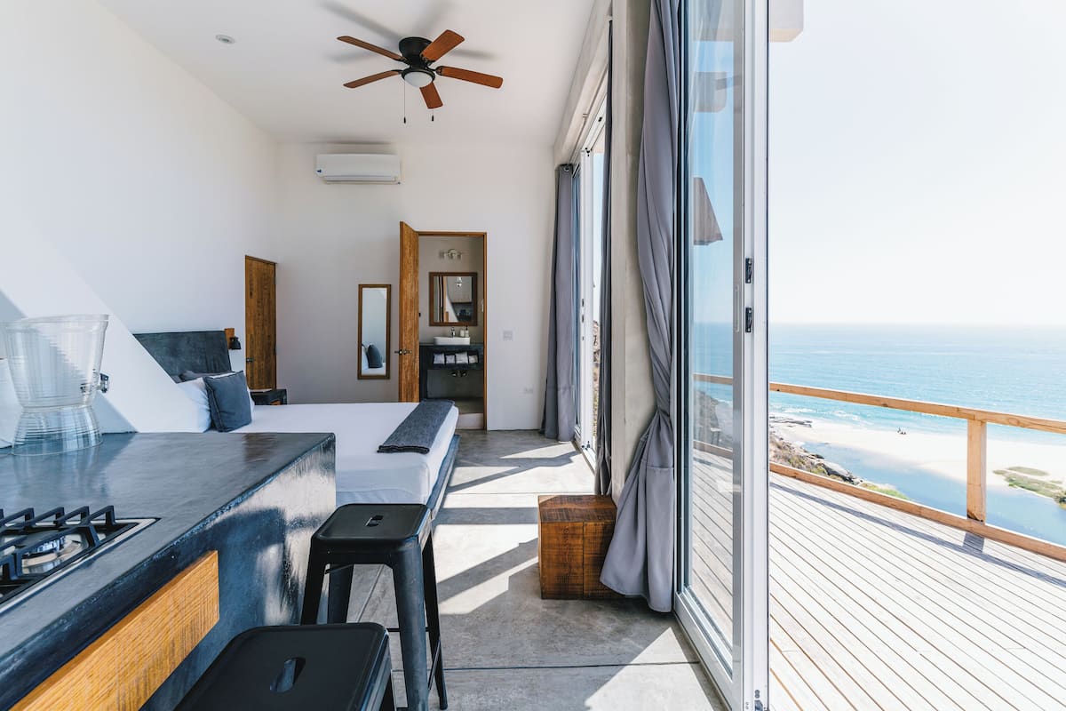 A modern bedroom with a balcony overlooking the sea, a wooden ceiling fan, and a large window with curtains.