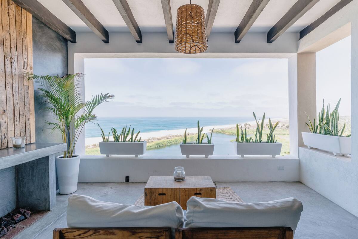 A modern room with a view of the ocean, featuring a wooden bench, a small table, and potted plants.