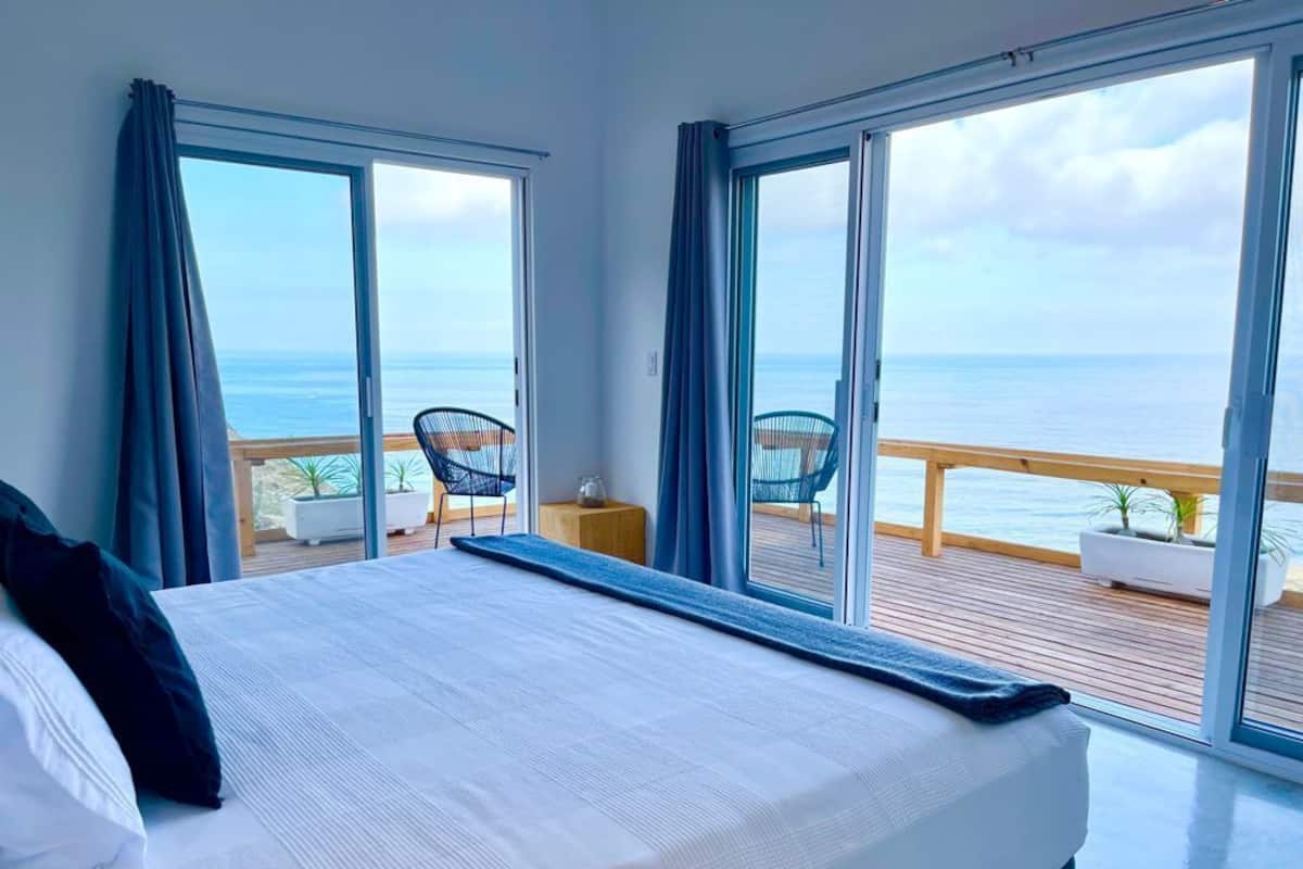 A bedroom with a large bed, a view of the ocean through floor-to-ceiling windows, and a balcony with outdoor seating.