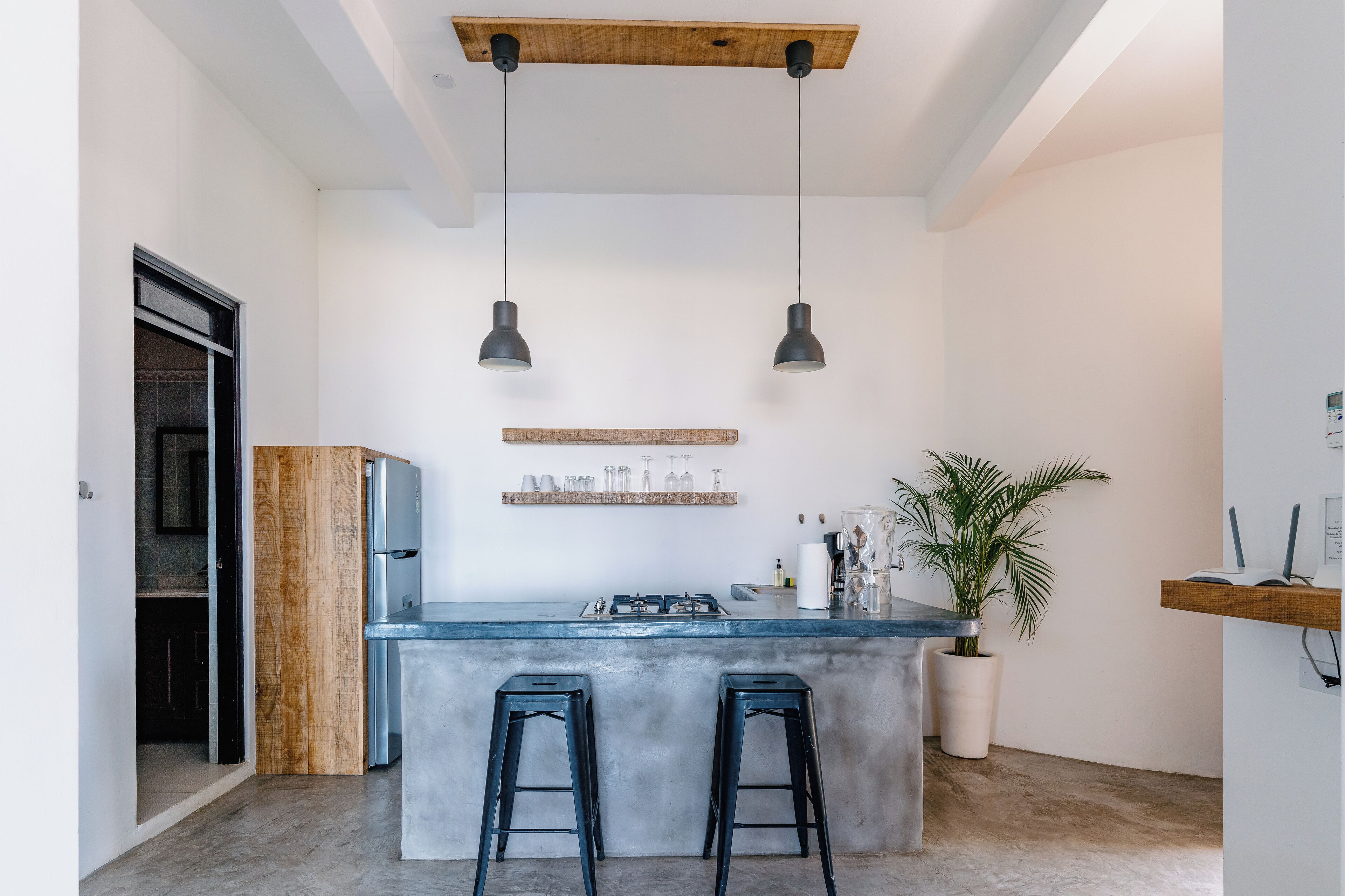 A modern kitchen with a central island, two bar stools, pendant lighting, a wooden shelf with glasses, and a potted plant.
