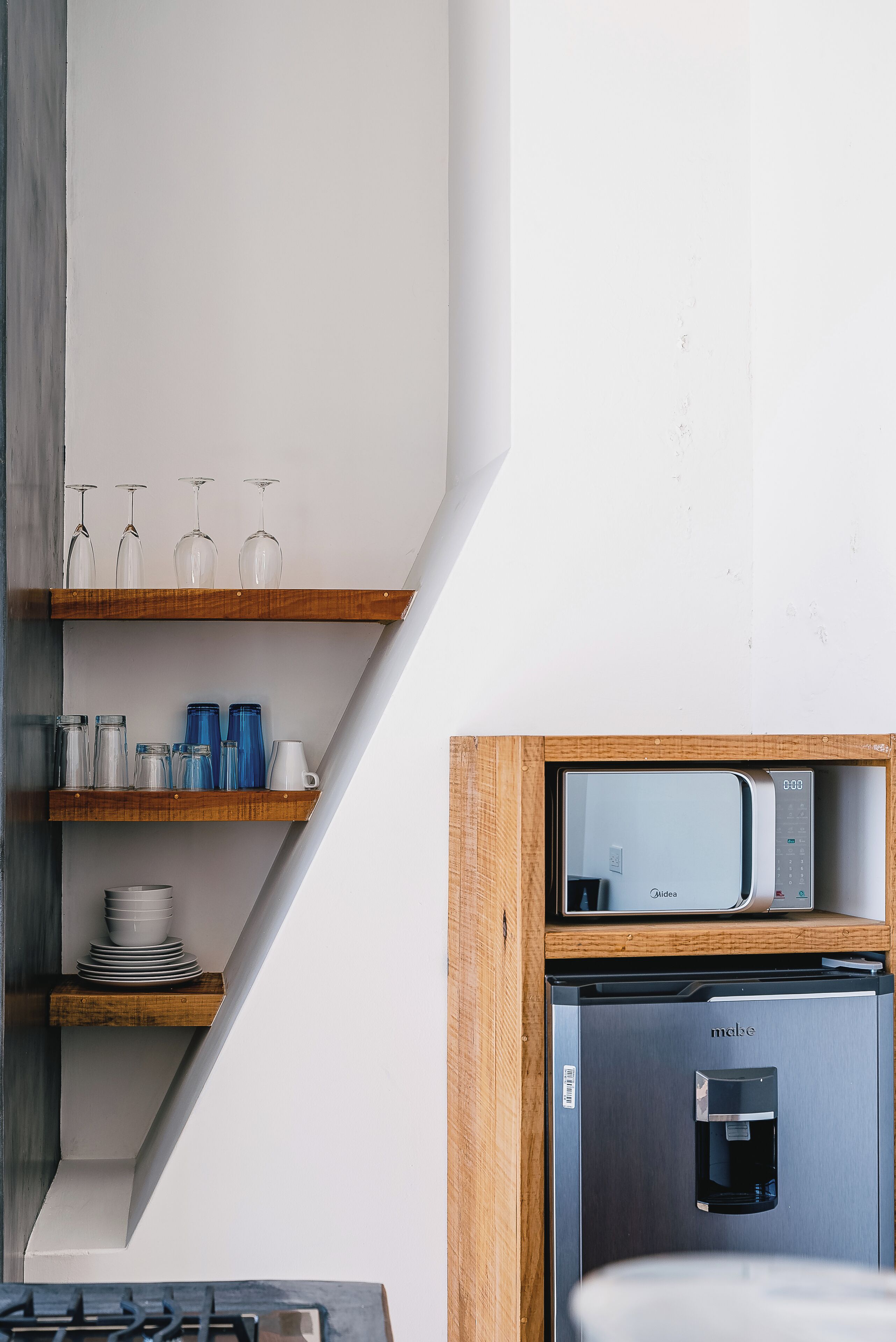 A kitchen with a built-in microwave and water dispenser, wooden shelves with glassware, and a stack of plates.